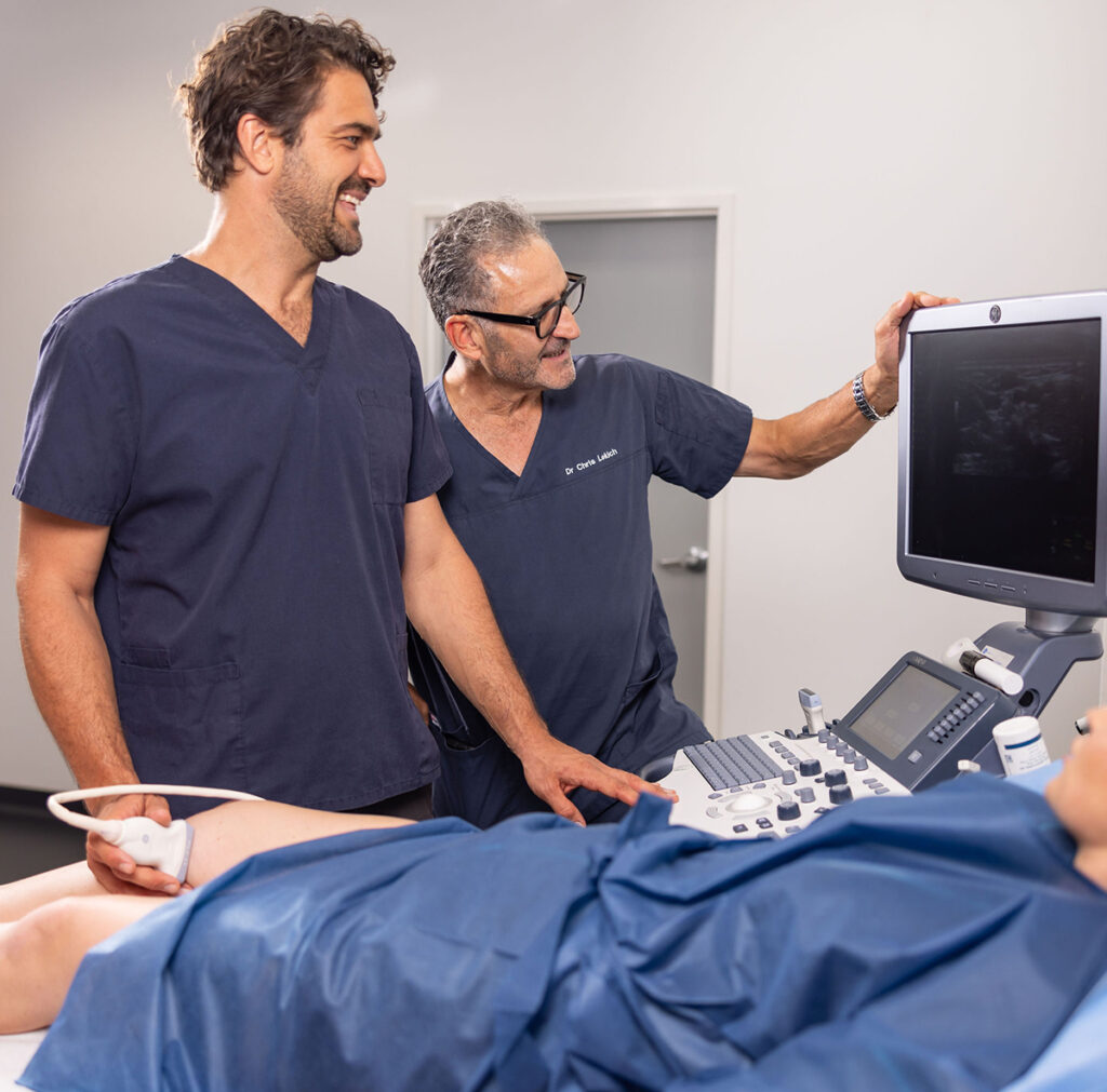 Two medical professionals examine an ultrasound machine while a patient rests on an exam table, covered with a blue sheet.